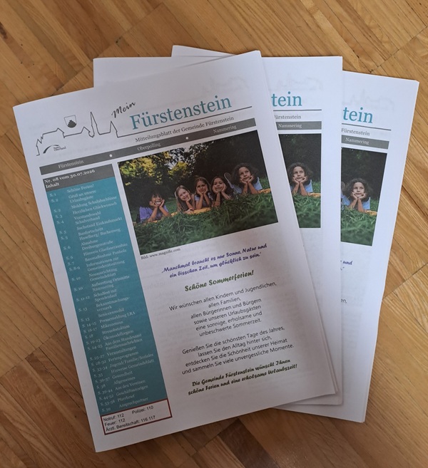Drei Ausgaben des Gemeindeblattes „Mein Fürstenstein“ liegen auf einem Holzboden gestapelt; auf dem obersten Exemplar ist auf dem Titelbild ein Foto von fünf lächelnden Kindern im Freien zu sehen, links davon ein Kalender. Der Text kündigt ein Sommerferienprogramm an.