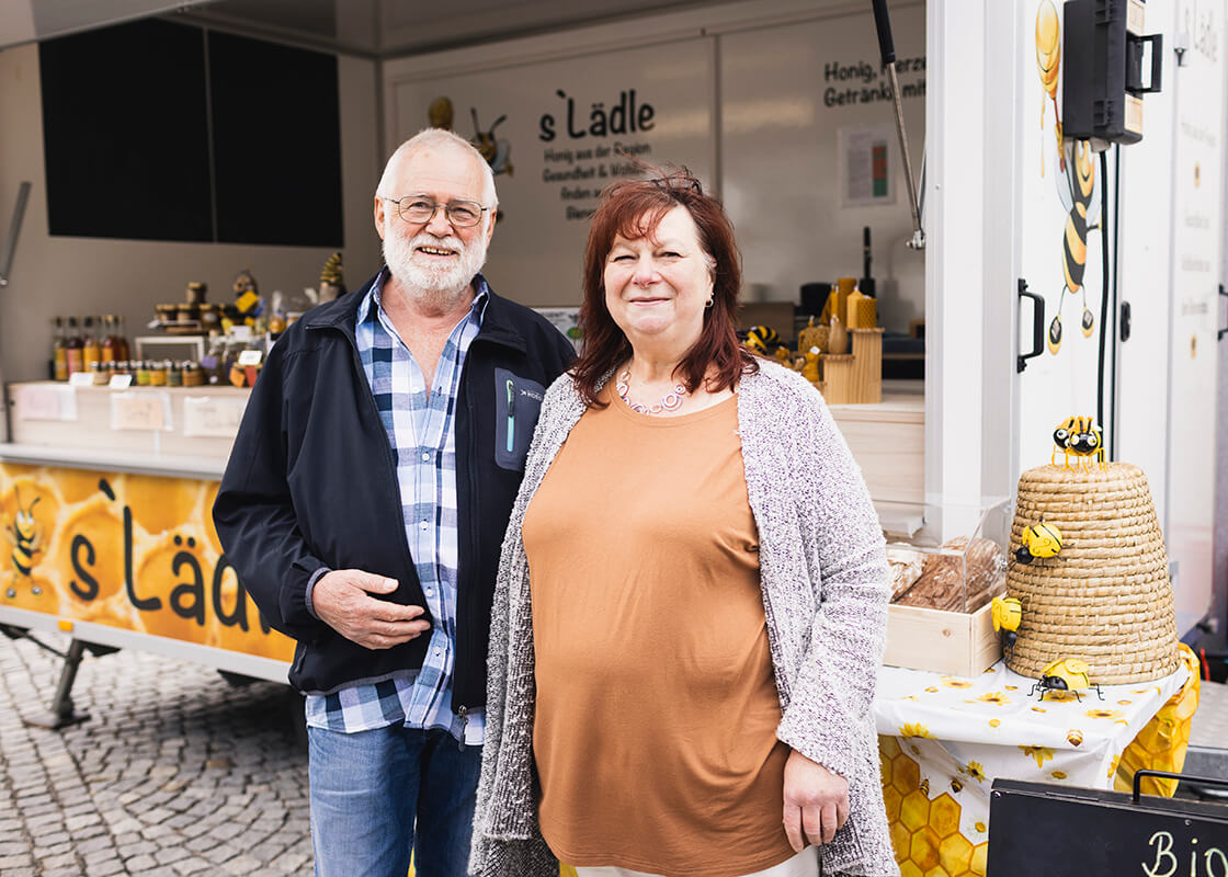 Ein älterer Mann und eine ältere Frau stehen vor einem Honigstand im Freien und lächeln. Hinter ihnen sind in Regalen Gläser mit Honig und verwandten Produkten ausgestellt. Der Stand ist gelb dekoriert und mit Artikeln zum Thema Bienen geschmückt.