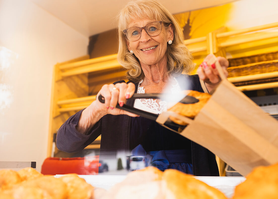 Eine ältere Frau mit Brille und einem strahlenden Lächeln legt mit einer Zange ein Gebäckstück in eine braune Papiertüte an der Theke einer Bäckerei, im Hintergrund sind Regale und Backwaren zu sehen.