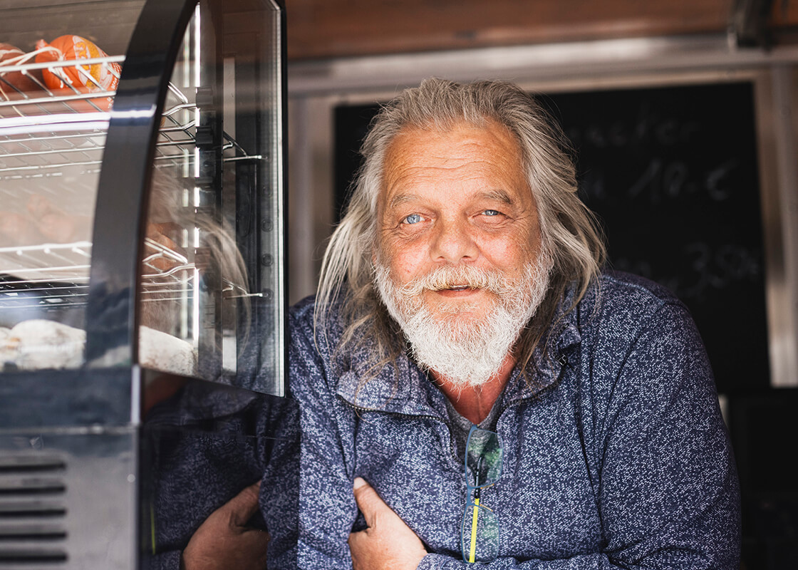 Ein Mann mit langen grauen Haaren und Bart, der einen blauen Pullover trägt, lächelt, während er sich an einer Theke neben einer Konditorei-Vitrine anlehnt; im Hintergrund ist eine Kreidetafel zu sehen.