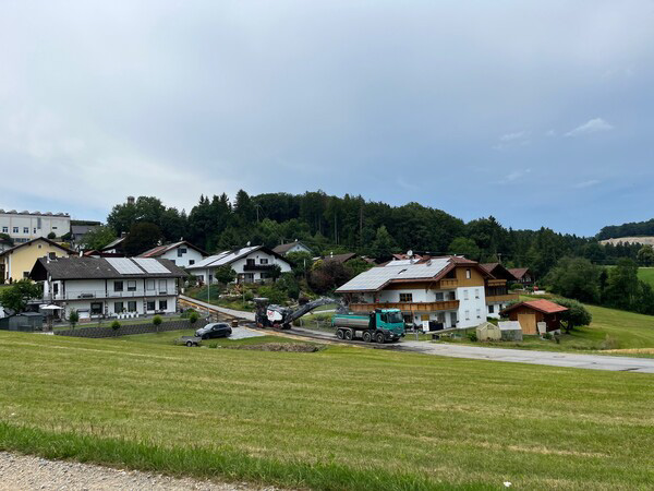 Straßenfräsarbeiten im Buchenweg in Fürstenstein. Eine Fräsmaschine und ein Lkw sind im Wohngebiet im Einsatz, im Hintergrund sind Wohnhäuser sowie die umliegende Landschaft zu sehen.