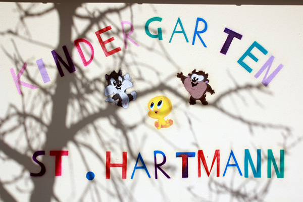 An einer Wand steht in bunten Buchstaben „KINDERGARTEN ST. HARTMANN“ geschrieben, wobei Comicfiguren und Schatten von Ästen den Text überlagern.