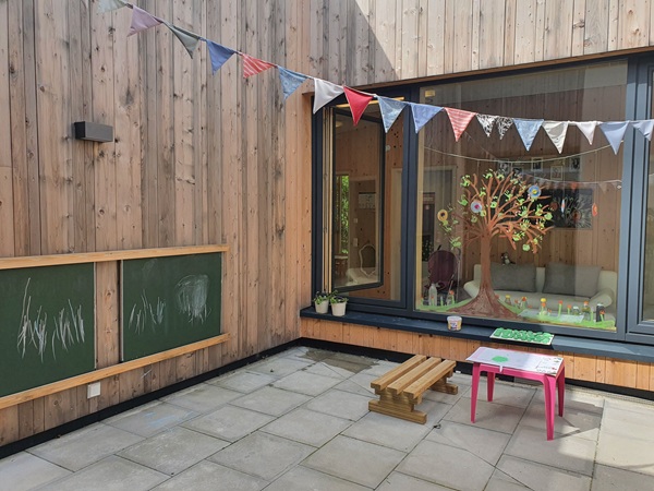 Innenhof der Kindertageseinrichtung Fürstenstein mit Kreativbereich, Sitzgelegenheiten und großen Fenstern zum Gruppenraum. Die naturnahe Gestaltung lädt zum Spielen, Lernen und Entdecken ein.