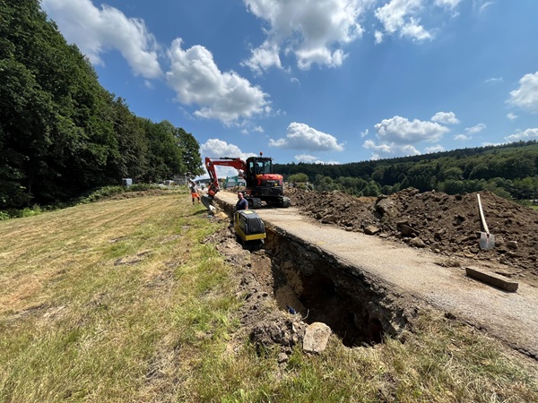 Bagger graben neben einer beschädigten Landstraße, in deren Nähe Arbeiter zu sehen sind. Die Straße ist teilweise eingestürzt, sodass Erde und Rohrleitungen freigelegt sind. Bäume und Wiesen umgeben die Baustelle unter einem blauen Himmel mit vereinzelten Wolken.