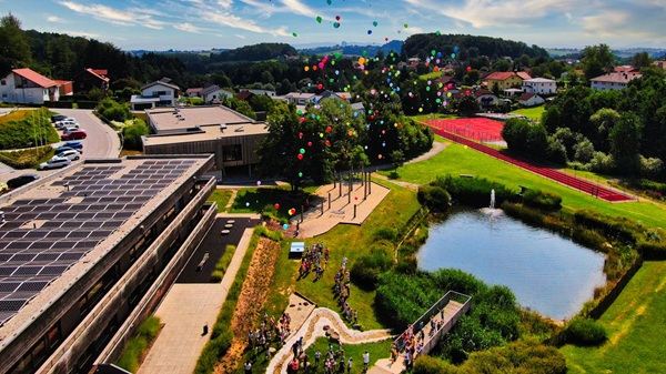 Luftaufnahme der Grundschule Fürstenstein mit Schulgebäude, Pausenhof, Schulgarten, Naturteich und Sportanlagen. Über dem Schulgelände steigen bunte Luftballons in den Himmel.