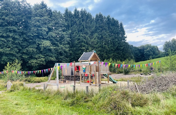 Ein Spielplatz aus Holz mit Schaukeln, Rutschen und einem Spielhaus ist mit bunten dreieckigen Wimpeln geschmückt. Der Spielplatz ist von Gras, Büschen und hohen Bäumen umgeben, über ihm erstreckt sich ein teilweise bewölkter Himmel.