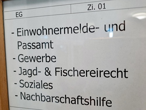 Auf einem Schild in deutscher Sprache sind folgende Ämter aufgeführt: „Einwohnermelde- und Passamt, Gewerbe, Jagd- und Fischereirecht, Soziales, Nachbarschaftshilfe.“ Es ist mit „EG“ und „Zi. 01“ beschriftet, was für Erdgeschoss, Zimmer 01 steht. Das Schild befindet sich im Innenbereich und ist mit Holz eingerahmt.