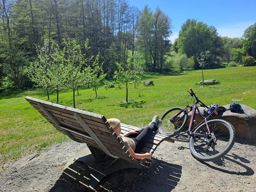 Eine Person entspannt sich auf einer Holzliege in einem sonnigen, grünen Park neben ihrem Fahrrad. Bäume und Wiesen umgeben die Gegend und schaffen eine friedliche Atmosphäre im Freien.