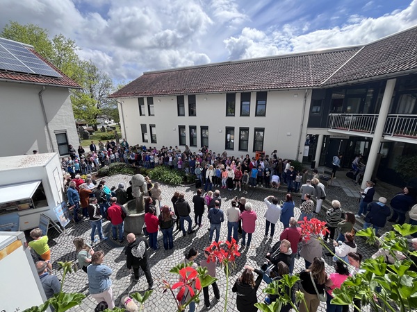 An einem teilweise bewölkten Tag versammelt sich eine große Gruppe von Menschen, darunter Kinder und Erwachsene, in einem Innenhof vor einem Gebäude mit rotem Ziegeldach. Einige stehen im Kreis, während andere zuschauen. Im Vordergrund sind leuchtend rote Blumen zu sehen.